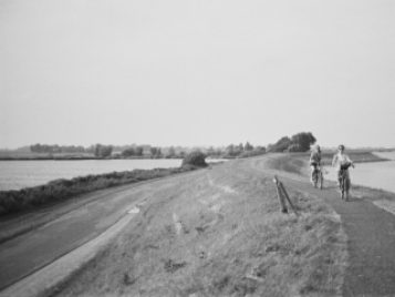 A black-and-white photograph of a curved trail that runs between two bodies of water. Two people are riding bicycles in the distance.